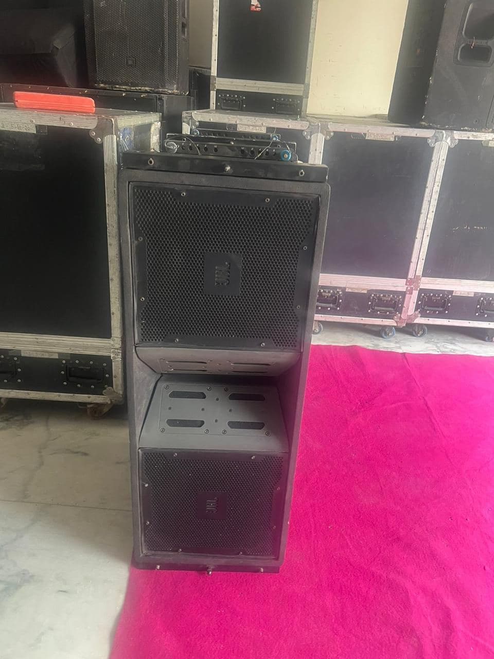 A JBL line-array cabinet standing on the venue carpet with flight cases stacked behind it
